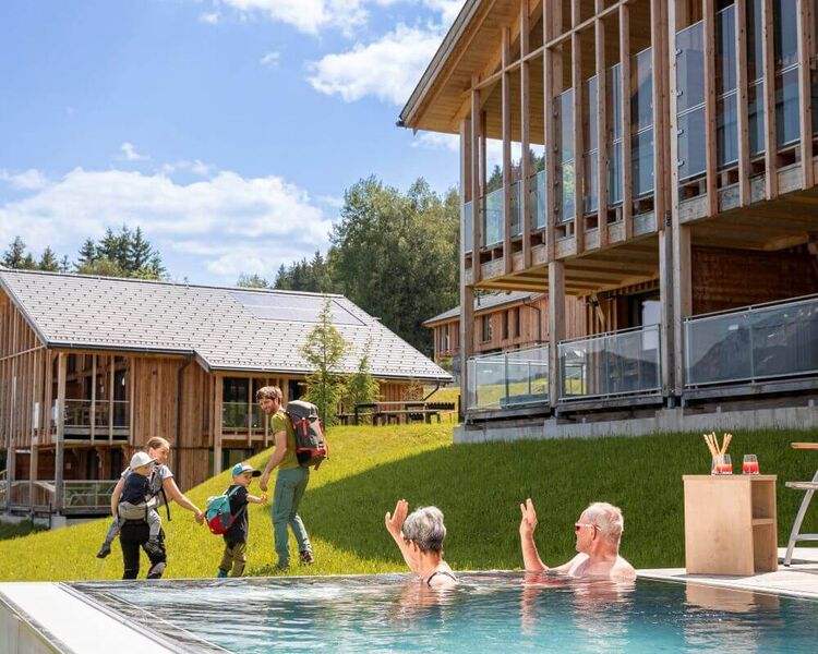 Großeltern winken der wandernden Jungfamilie aus dem privaten Pool ihres Ferienhauses zu.